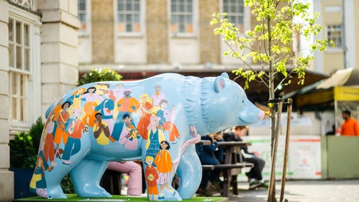 A finished blue bear decorated with people stands bold in the streets. A building and tree in the background.